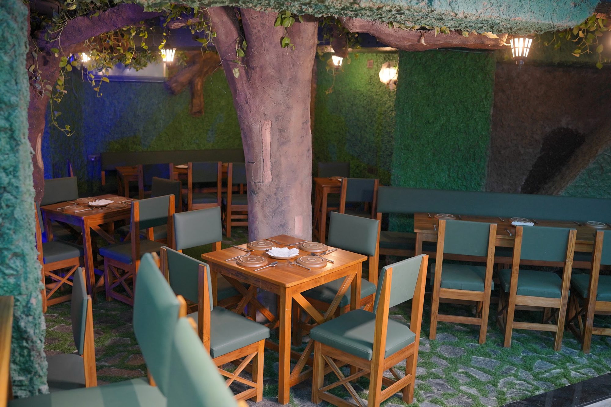 Dining tables under a giant tree with lanterns and moss walls at Forest Rogue Brews
